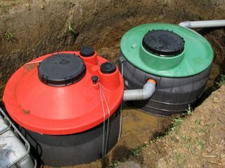 Two plastic septic tanks with red and green lids installed in a dirt trench, connected by a grey pipe.