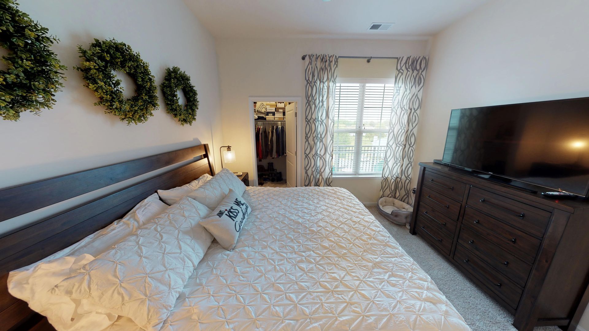 A bedroom with a large bed , dresser , television and wreaths on the wall.