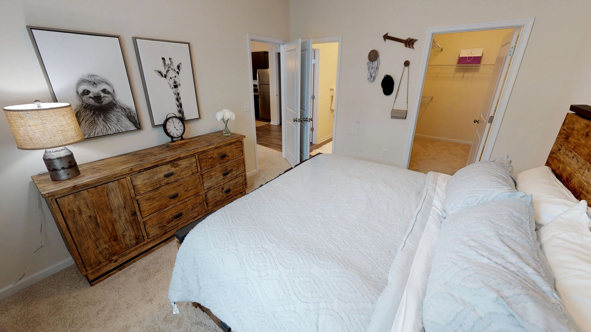 A bedroom with a bed , dresser , lamp and paintings on the wall.