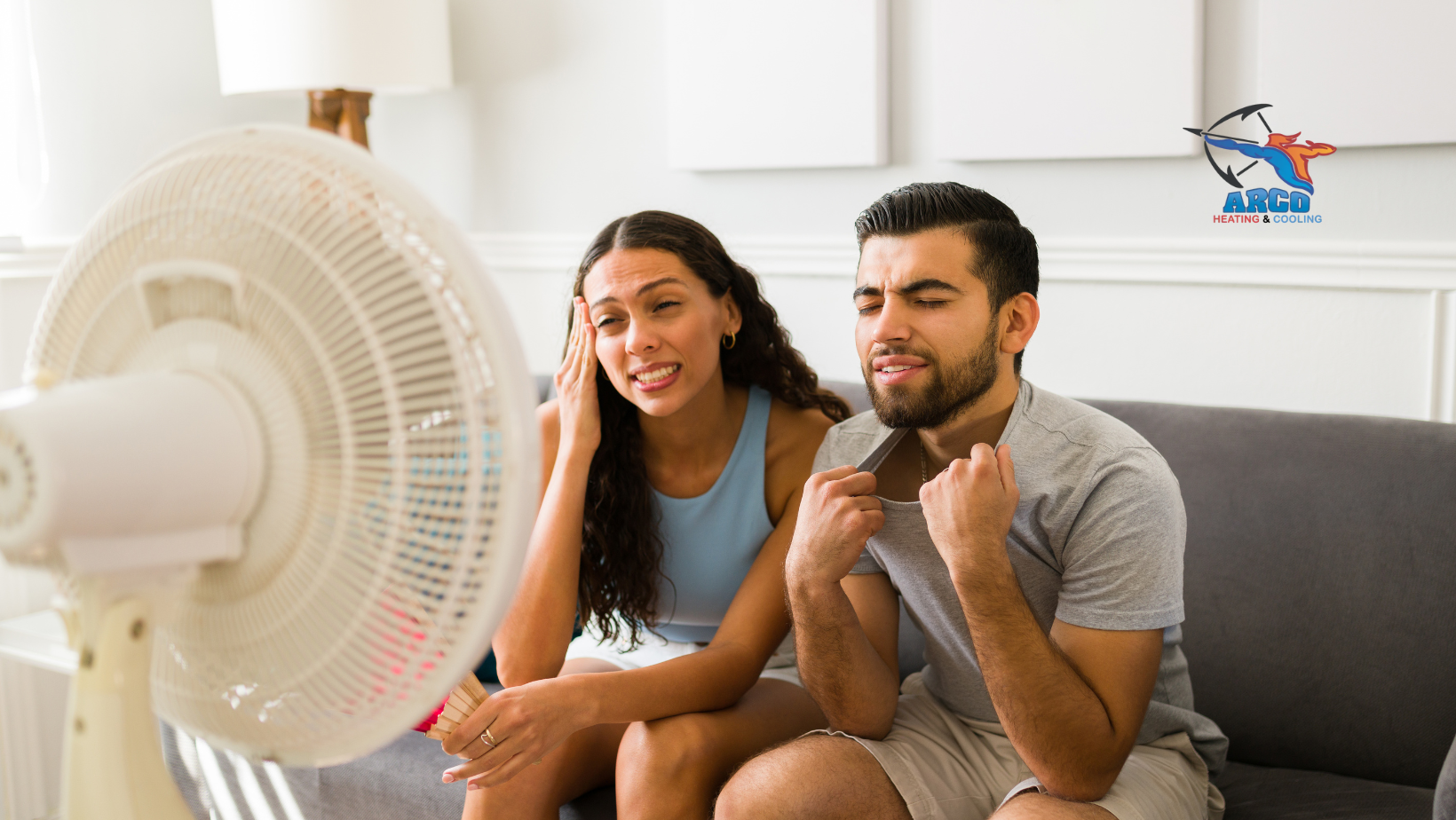 A man and a woman are sitting on a couch in front of a fan.