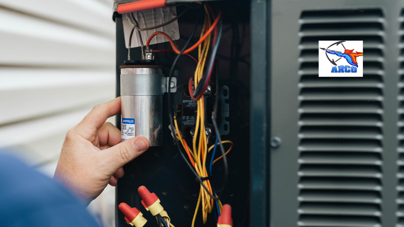 Hand holding an HVAC capacitor in an open unit, with wires, connectors, and a ARCO logo.