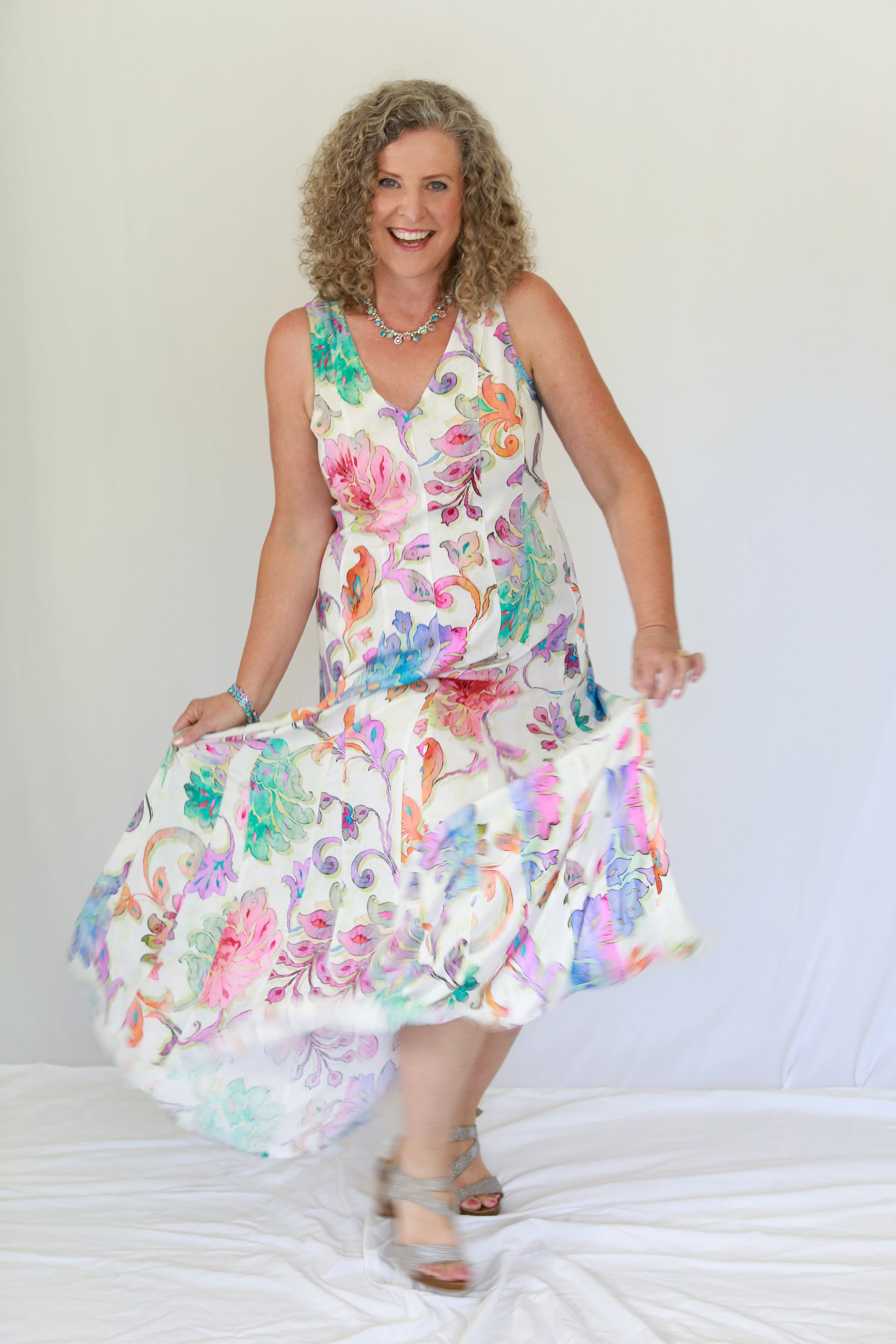 Woman in floral dress twirling, smiling. Wearing a necklace, heels, standing on white surface.