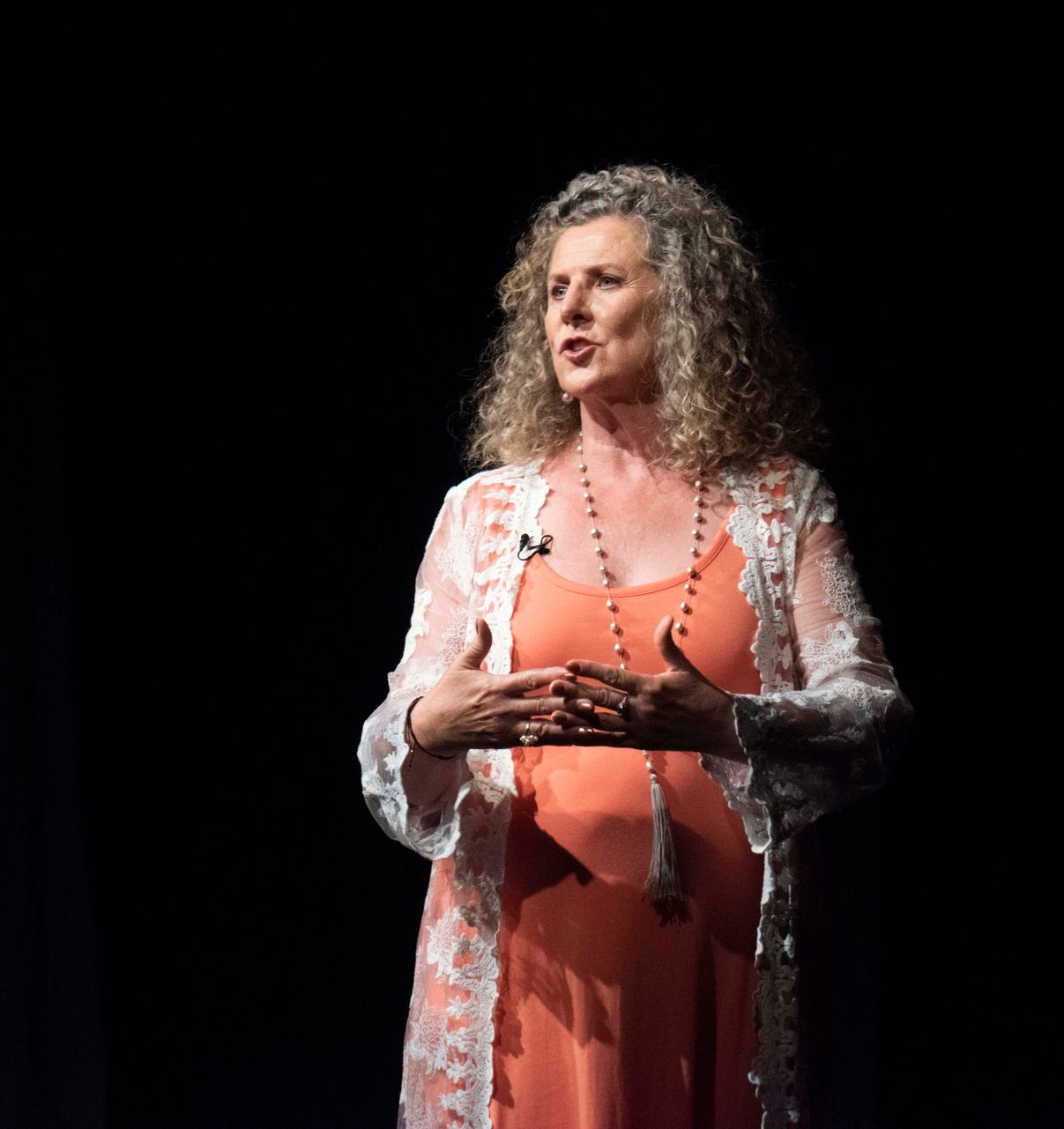 Woman on stage, speaking with hands gesturing, wearing orange dress and lace cardigan. Black background.