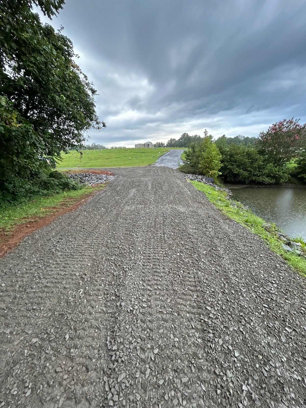 A gravel road leads uphill under a cloudy sky, with a pond to the right and green fields in the distance.