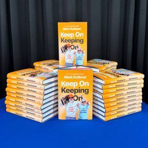 Stacks of "Keep On Keeping On" books with an orange cover, on a blue table.