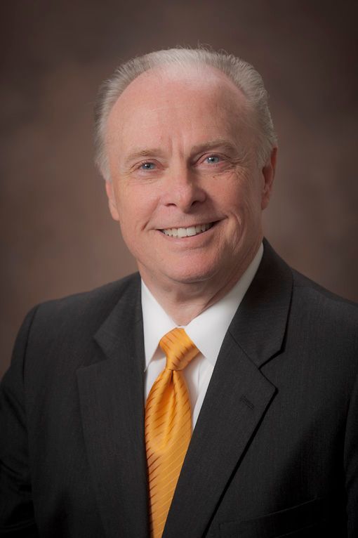 Dr. Mark Rutland in suit smiles, orange tie, against a brown gradient background.