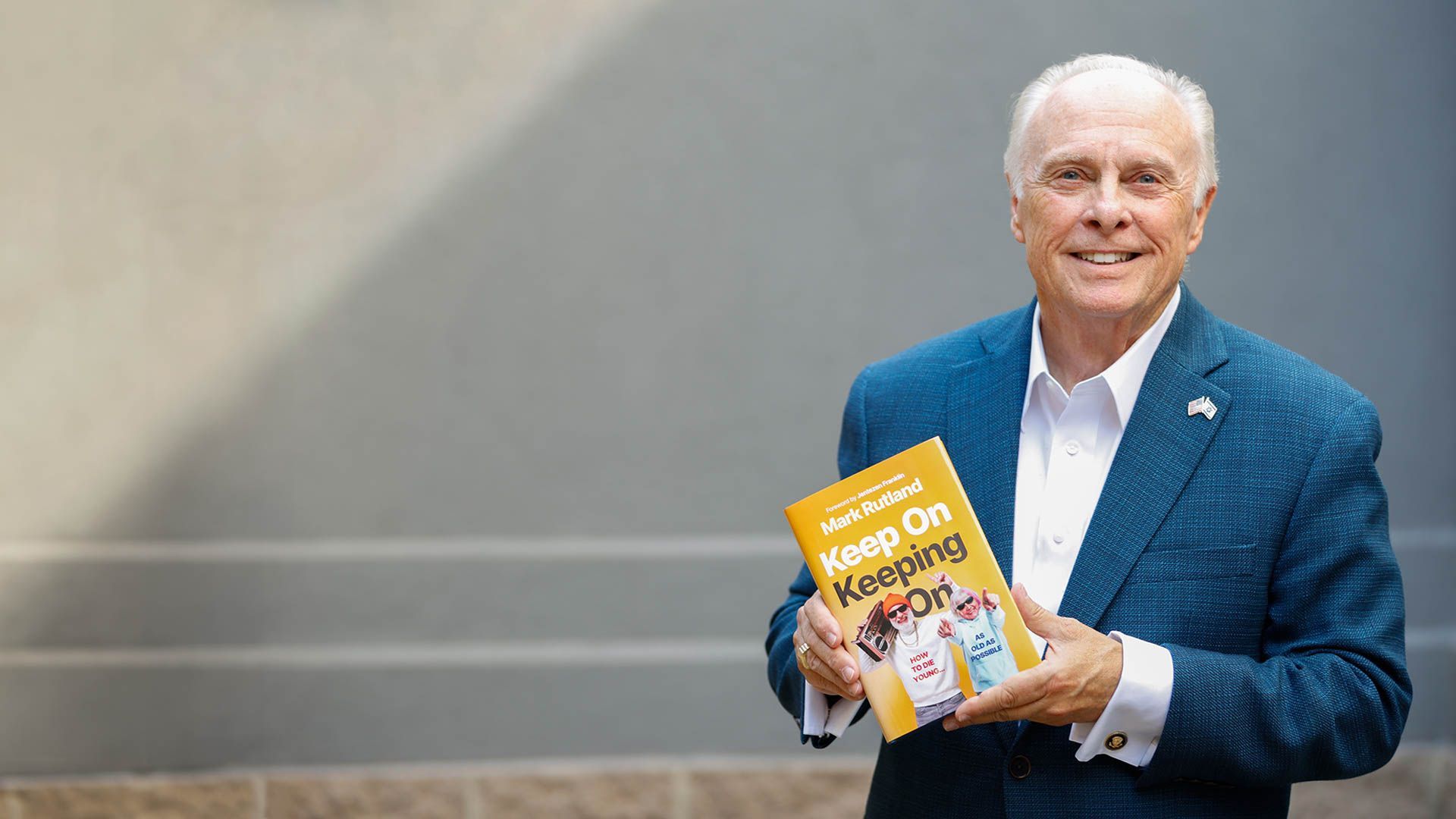 Dr. Mark Rutland holding a book titled "Keep On Keeping On" smiles, wearing a blue blazer, standing outdoors.