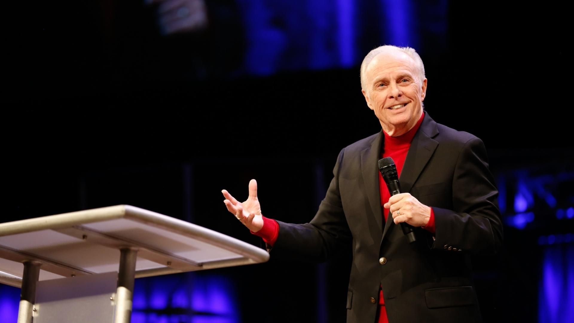 Dr. Mark Rutland at a podium, gesturing with his hands. He wears a black suit, red shirt, and is on a stage.