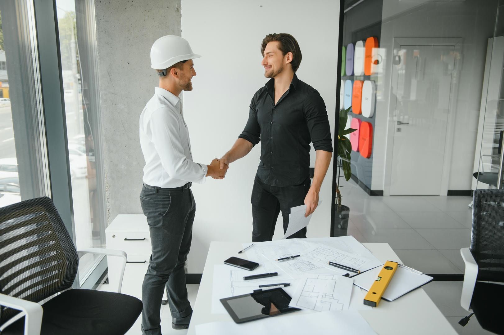 Two men are shaking hands in an office while wearing hard hats.