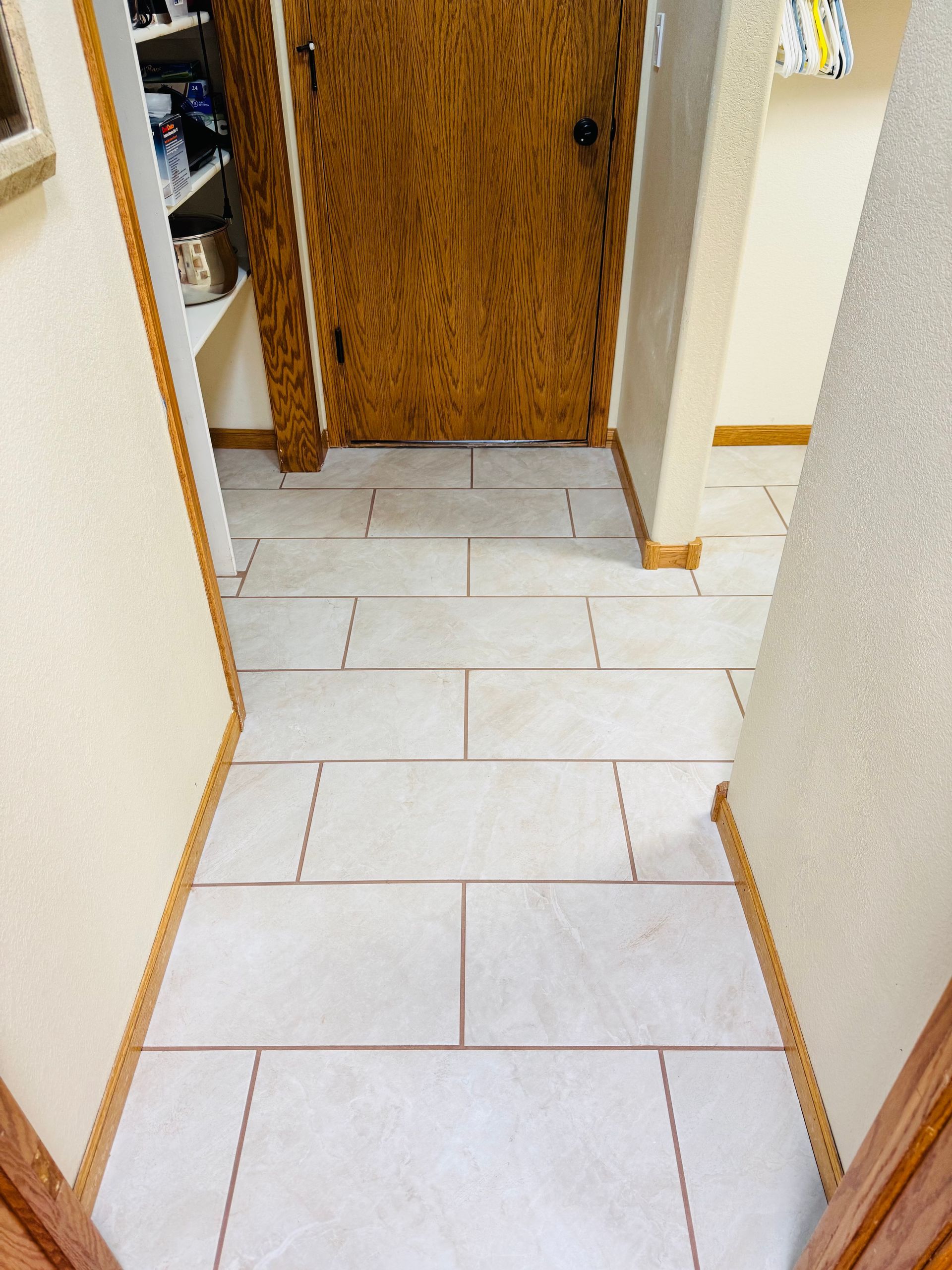 Hallway with rectangular light-colored tiles, brown grout lines, and wooden door.
