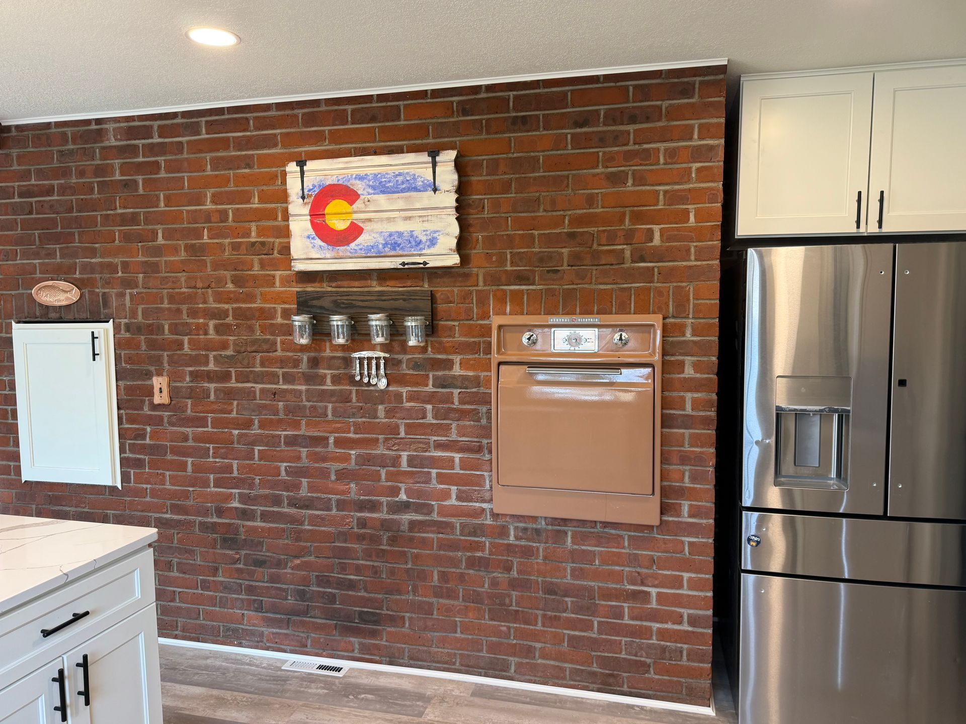 Brick wall kitchen with Colorado sign, oven, and stainless steel refrigerator. White cabinets on the right.