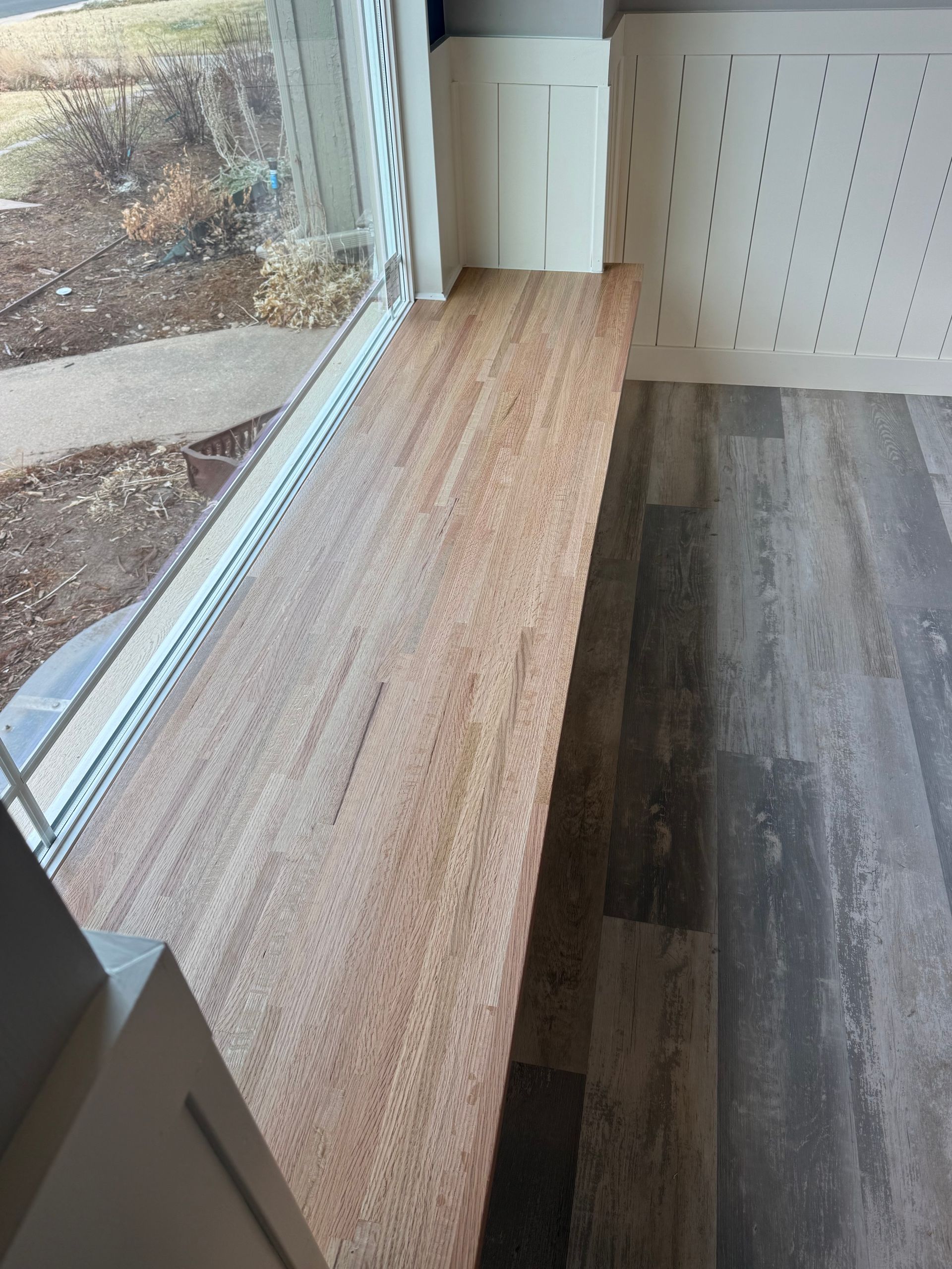 Wood-plank bench under a window, next to vinyl flooring. White wall with small holes is visible.