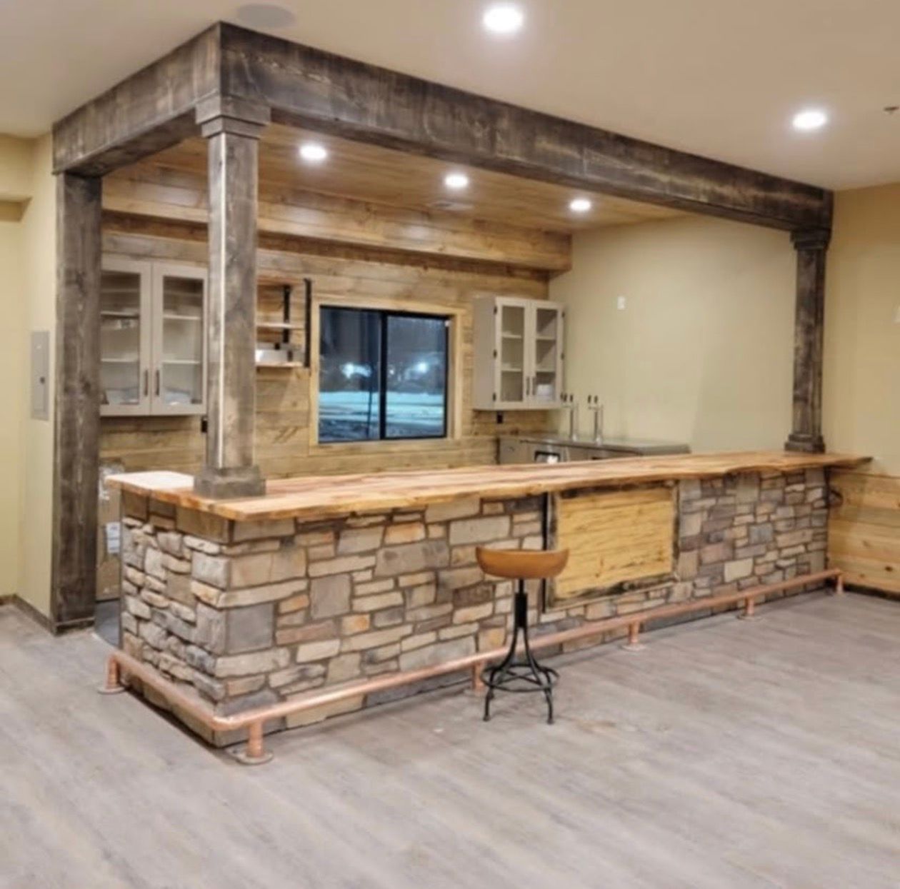 Rustic stone bar with wooden accents, copper footrest, and a stool in a finished basement.