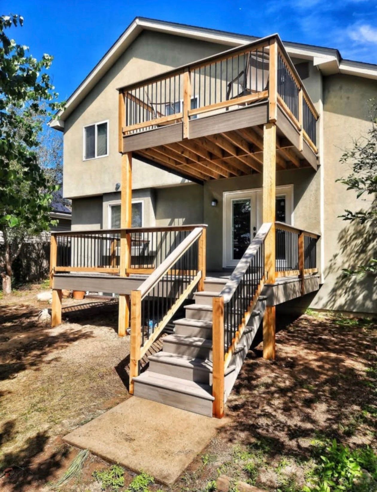 Two-story wooden deck attached to a house with stairs. Brown deck, grey stairs, tan siding, and trees.