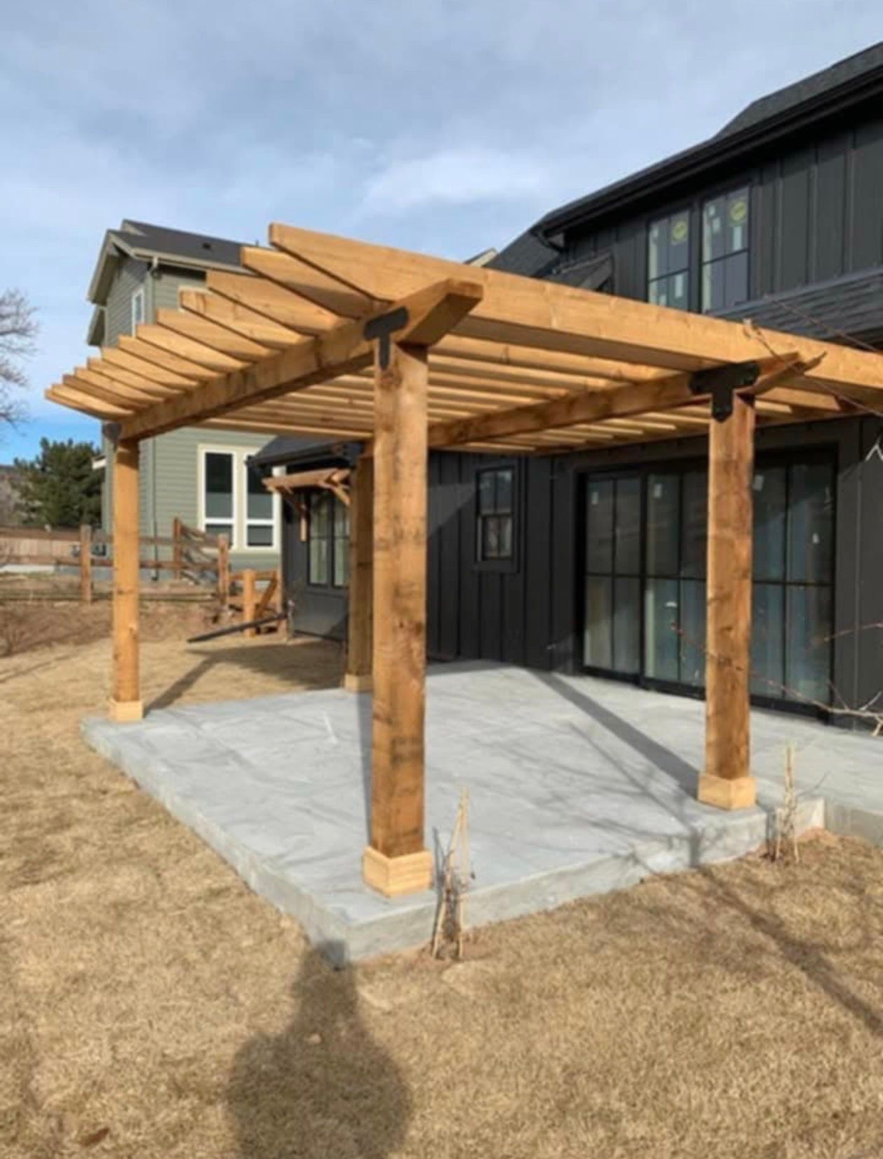 Wooden pergola over a concrete patio outside a dark-colored house on a sunny day.