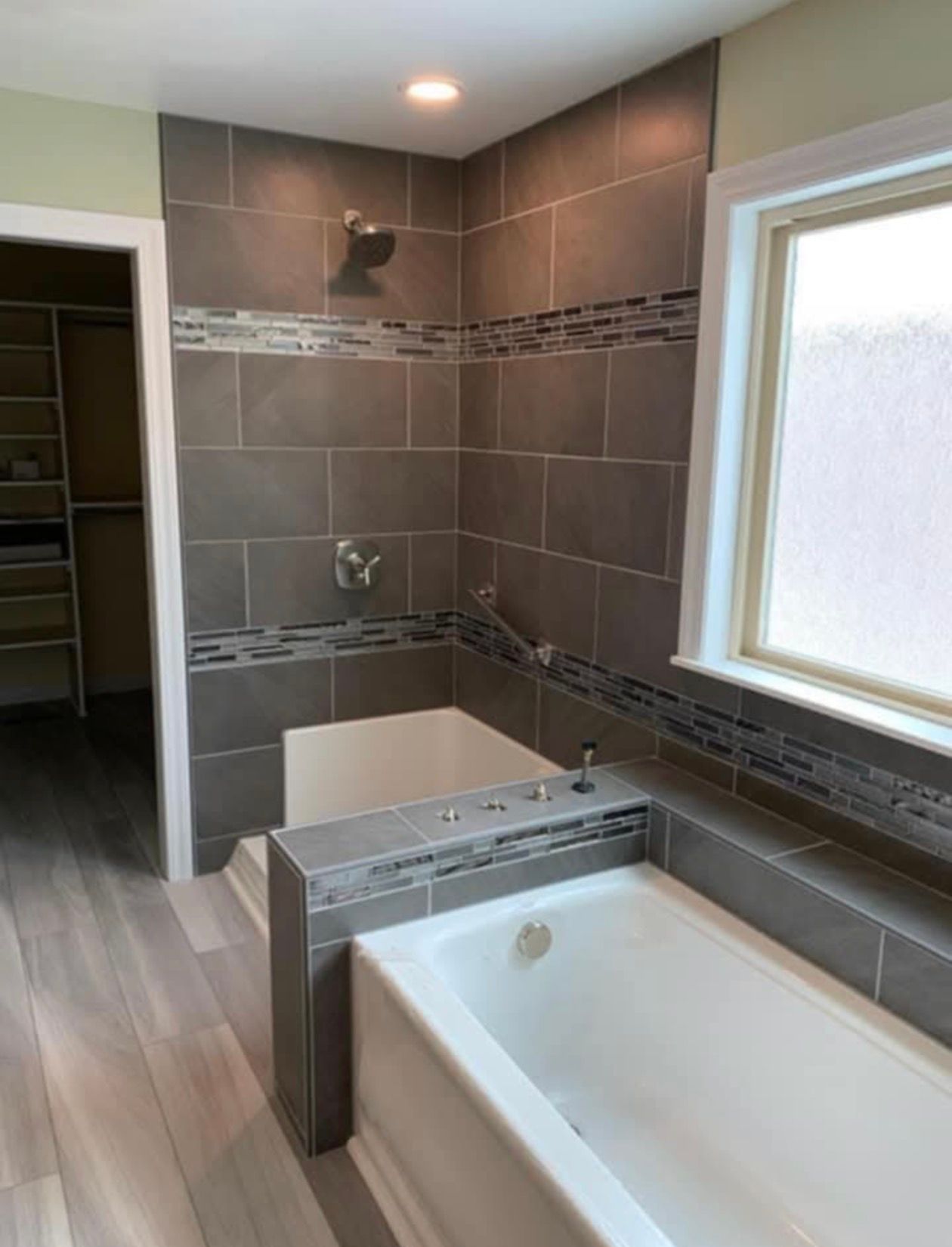 Bathroom with a tiled shower, bathtub, and a window. Gray tile and light wood flooring.