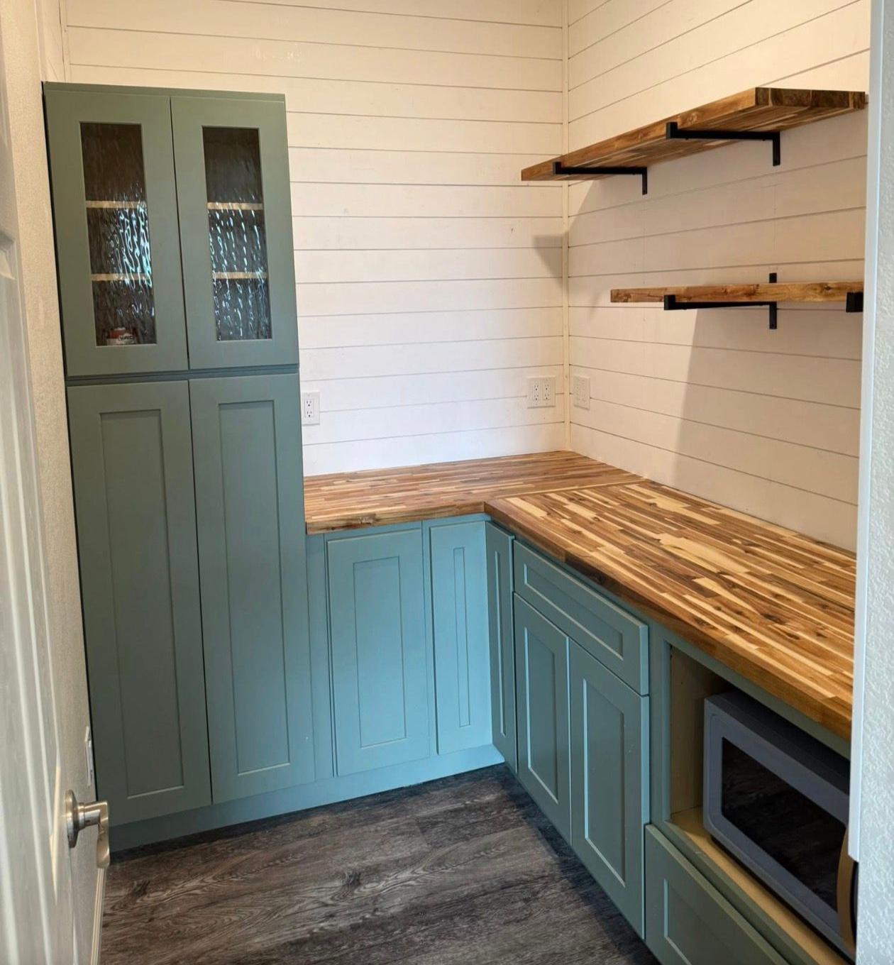 Small pantry with teal cabinets, wood countertops, and white shiplap walls. Open shelves and microwave.