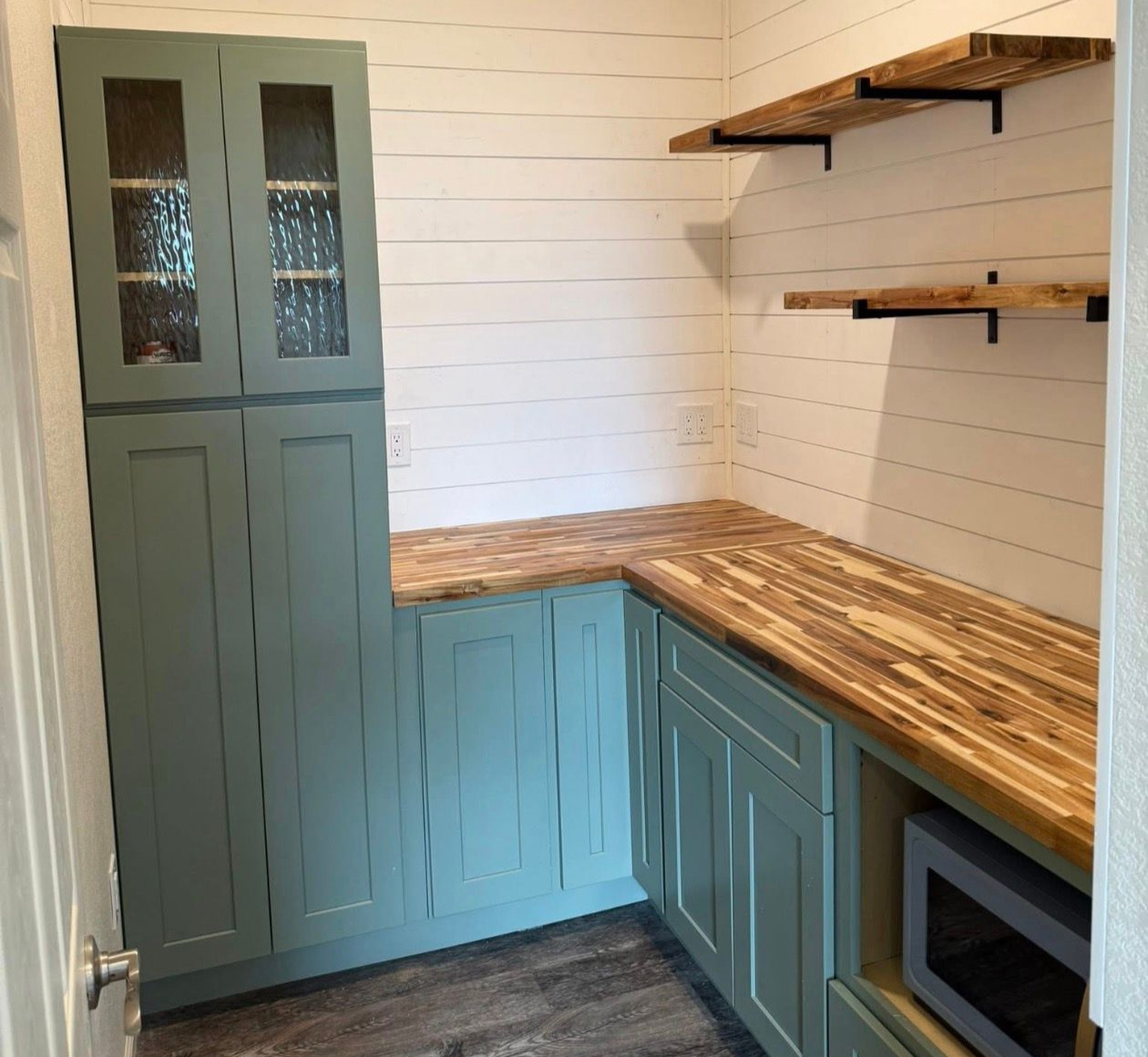 A small pantry with green cabinets, butcher block countertops, white shiplap walls, and wood shelves.