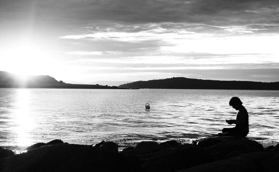 A black and white photo of a person sitting on a rock near the water