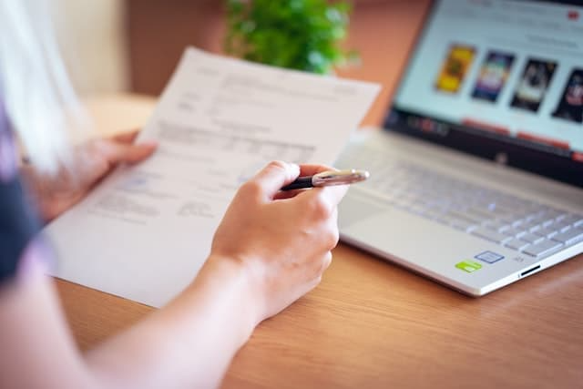 A person is holding a pen and a piece of paper in front of a laptop computer.