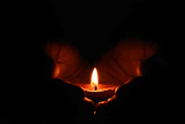 A person is holding a lit candle in their hands in the dark.