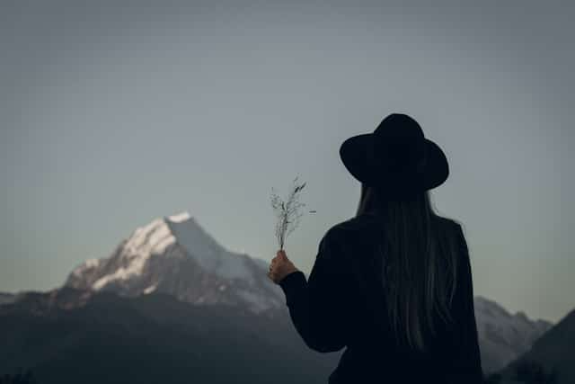 A woman in a hat is holding a flower in front of a mountain.