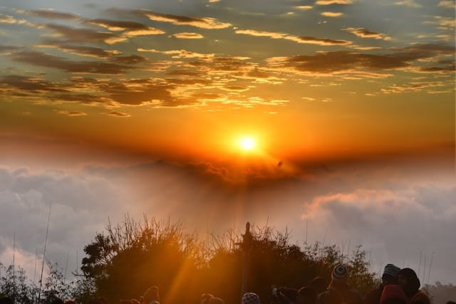 A group of people are standing on top of a hill watching the sun set over the clouds.