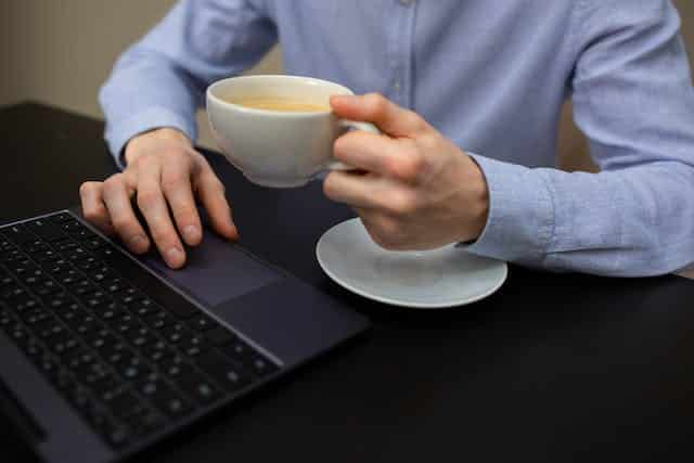 A man is holding a cup of coffee while using a laptop computer.