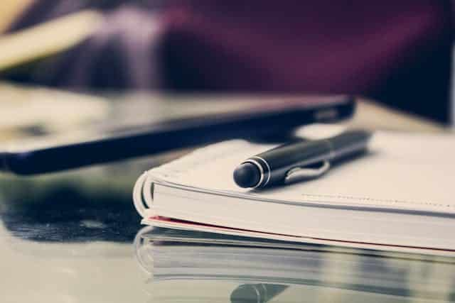 A pen is sitting on top of a notebook next to a cell phone.