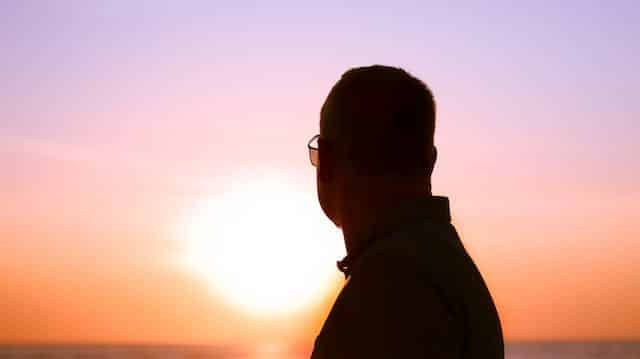 A man wearing glasses is standing on a beach at sunset.