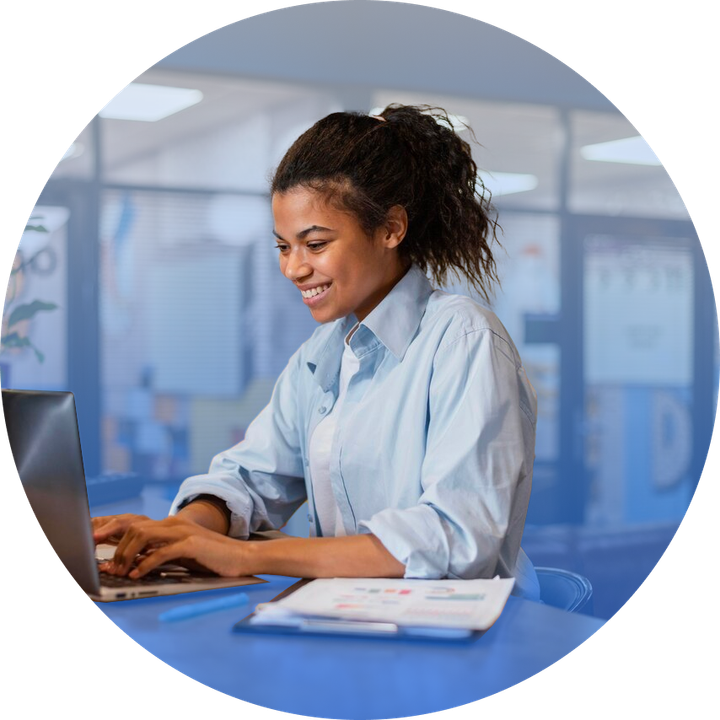 Woman with dark skin, ponytail, smiling, typing on laptop at blue desk. Papers and pen are on desk.