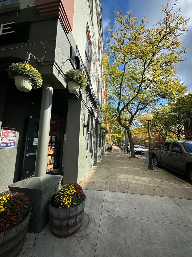 Sidewalk of shops with hanging flower baskets, trees, and a truck parked along a city street.