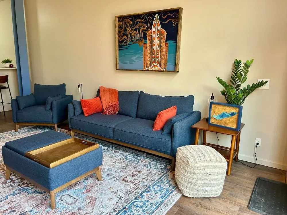 Living room with blue sofa, chairs, and rug; orange pillows; art on wall.