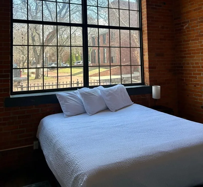 A bed with white bedding sits beneath a large window in a brick-walled room, looking outside.