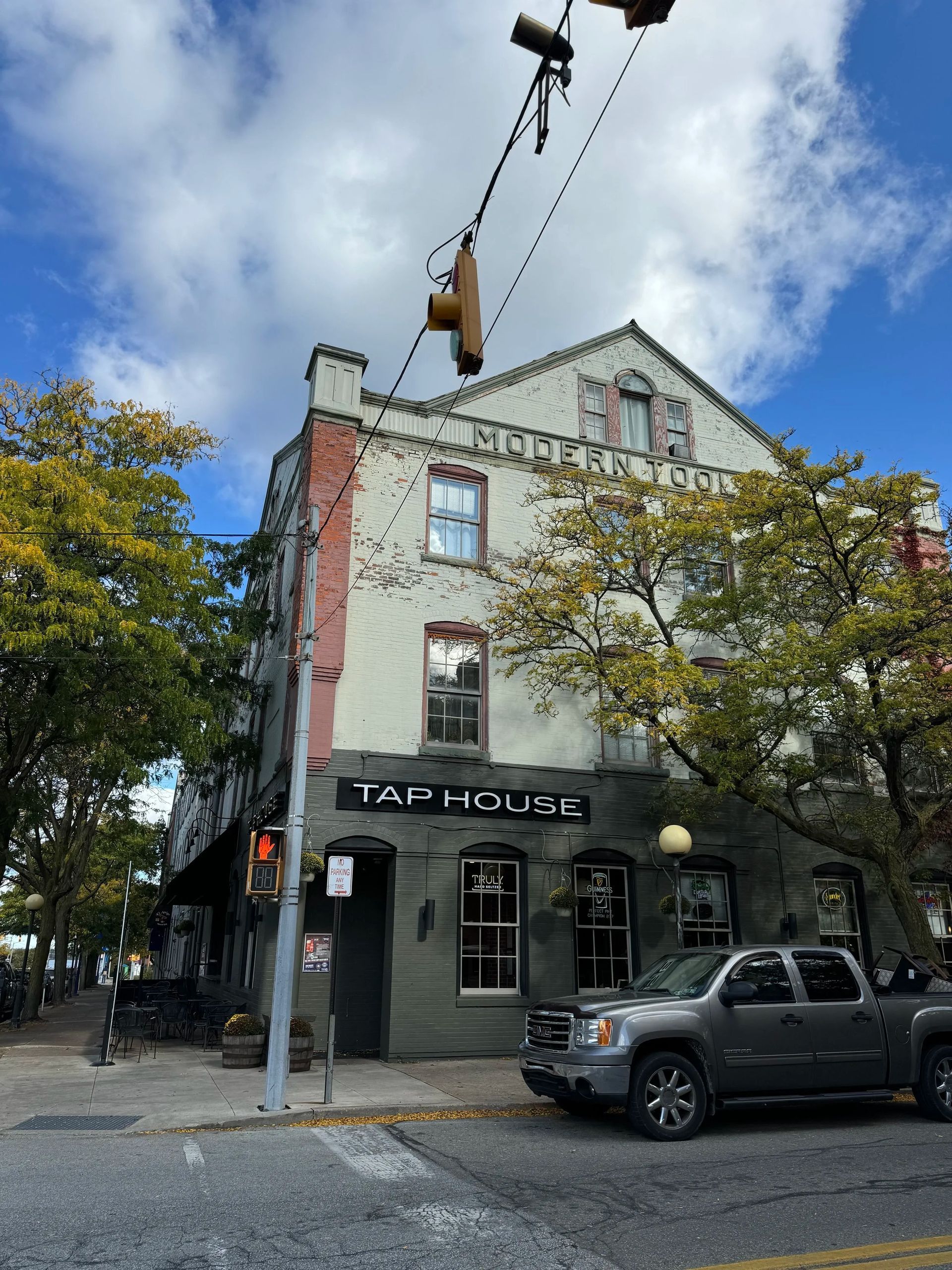 Taphouse building on a corner. Gray stucco with red brick, green door, sign. Cloudy sky and a silver truck parked.