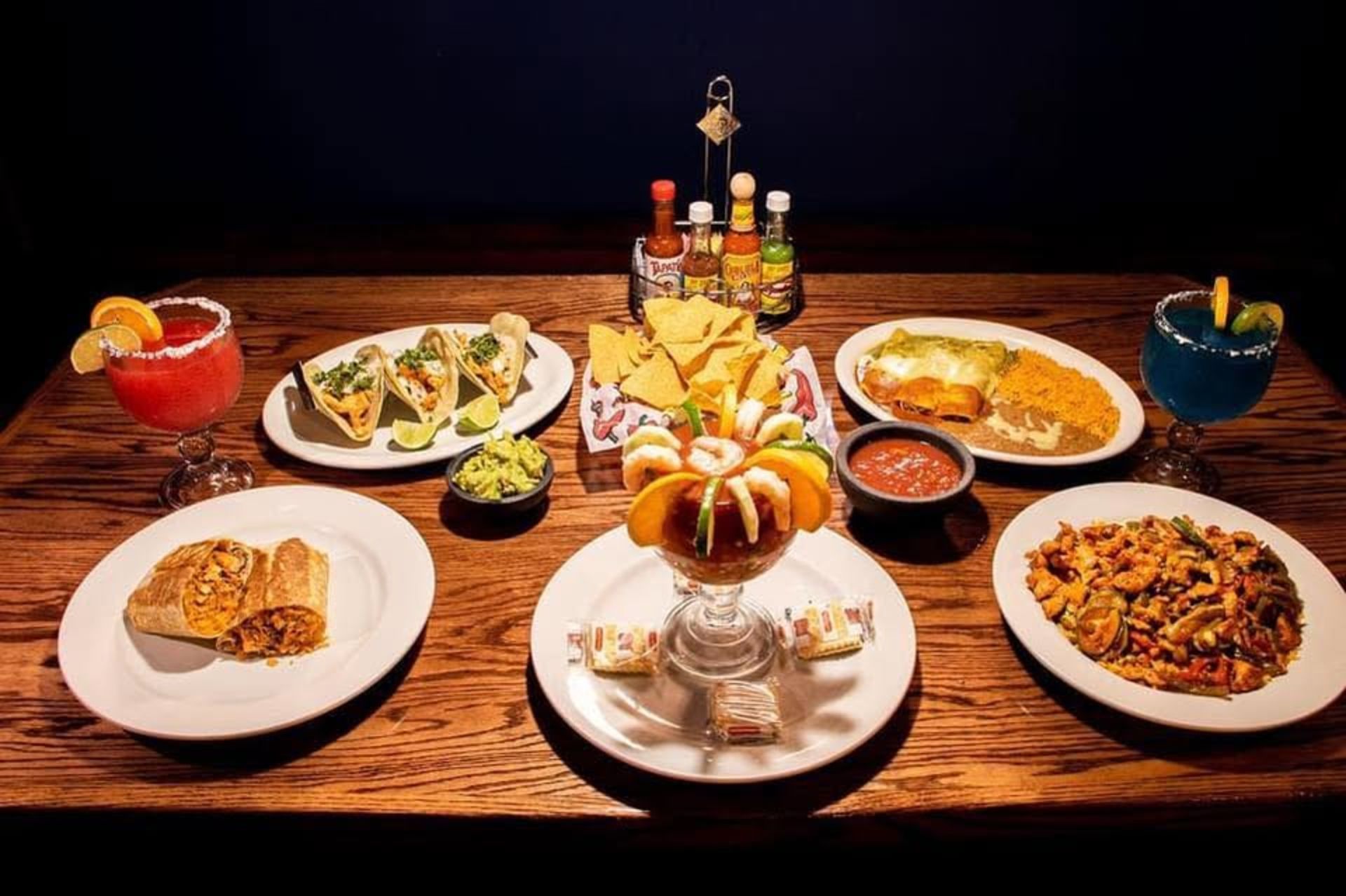 Mexican food spread on a wood table: tacos, burrito, shrimp dish, cocktails, guacamole, shrimp cocktail, and salsa.