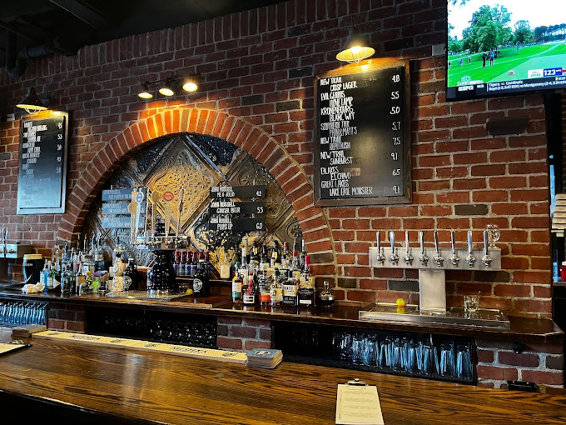 Brick-walled bar interior with dark wooden bar, beer taps, menu boards, and a TV showing sports.