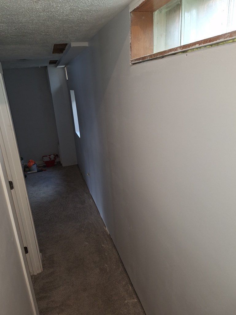 A narrow hallway with grey walls, a grey carpet, and an unfinished window frame near the ceiling.