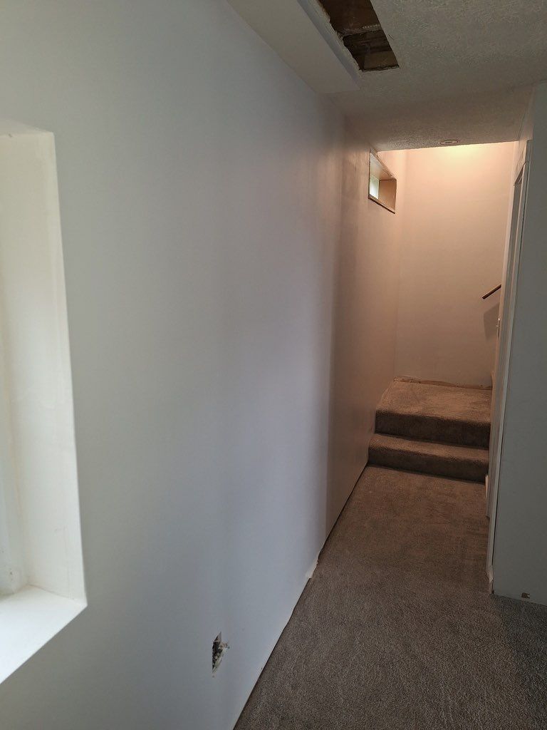 A perspective view of a carpeted hallway leading to short stairs, with white walls and an unfinished ceiling patch.