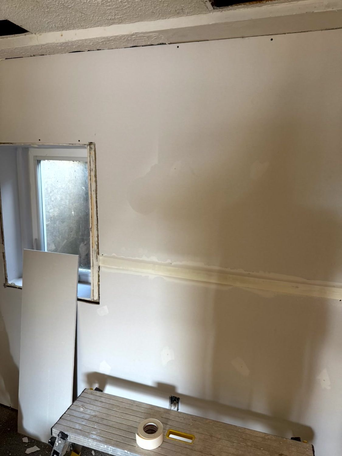 A windowed interior wall undergoing drywall repair with taped seams, showing a work platform with tape and tools below.