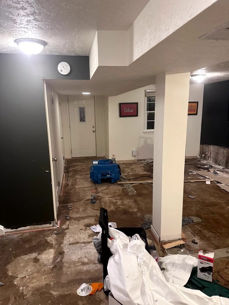 An indoor area undergoing water damage restoration, featuring exposed flooring, a blue industrial fan, and white walls.