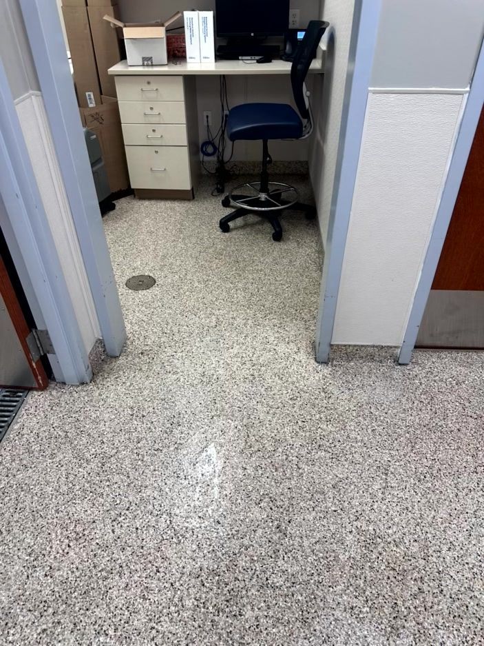 A view looking into a small office cubicle featuring a desk with a computer, a blue rolling chair, and tiled flooring.