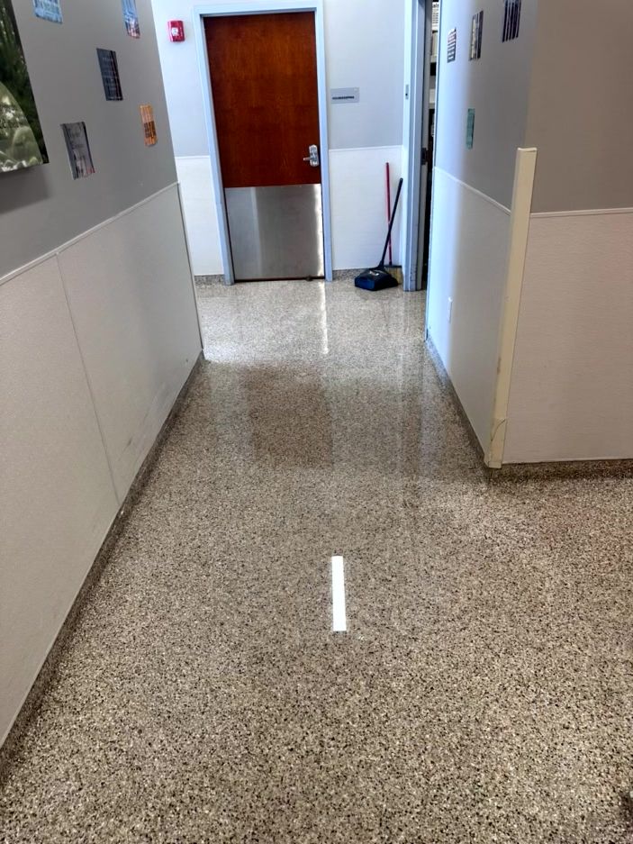 A brightly lit hallway with a shiny, speckled floor, leading to a closed wooden door with a metal kick plate.
