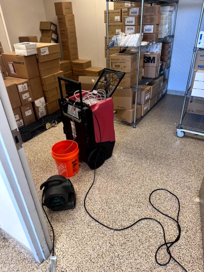 A portable dehumidifier and a small fan in a supply room with stacked cardboard boxes on metal shelving units.