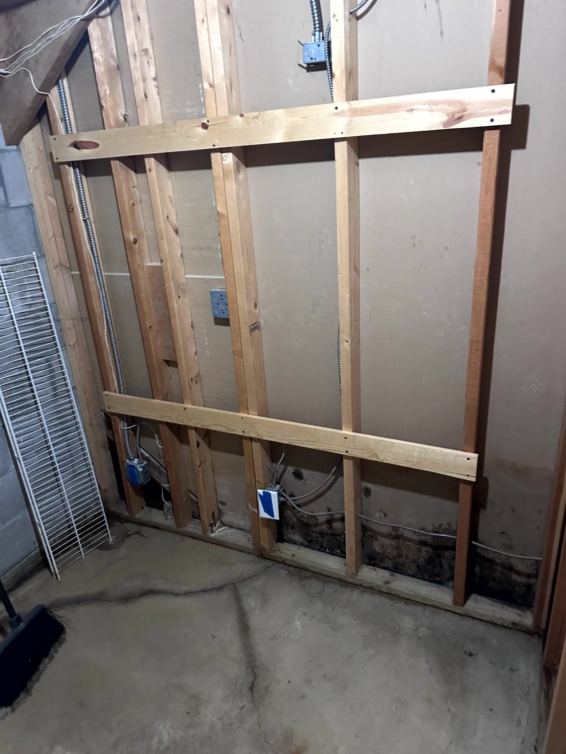 An unfinished basement wall frame made of wood studs, featuring electrical boxes and visible water stains at the base.