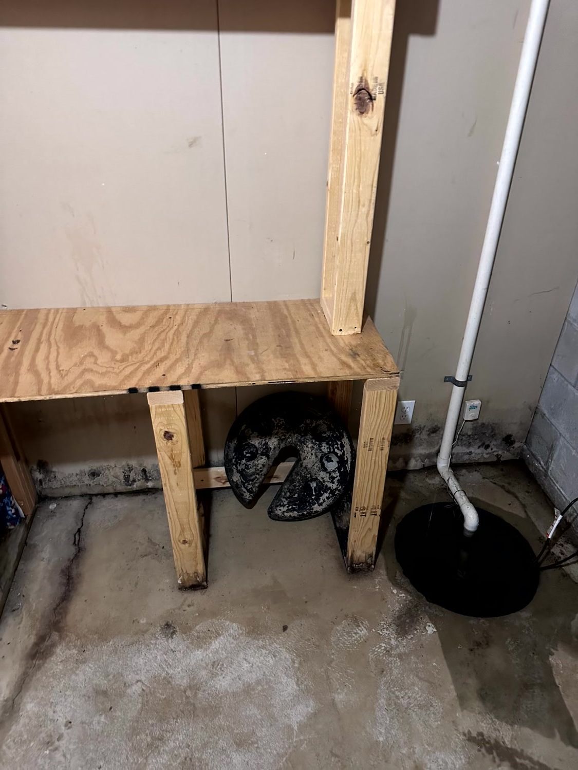 A wooden workbench in a basement, featuring a sump pump basin and pipe to the right and a circular object underneath.