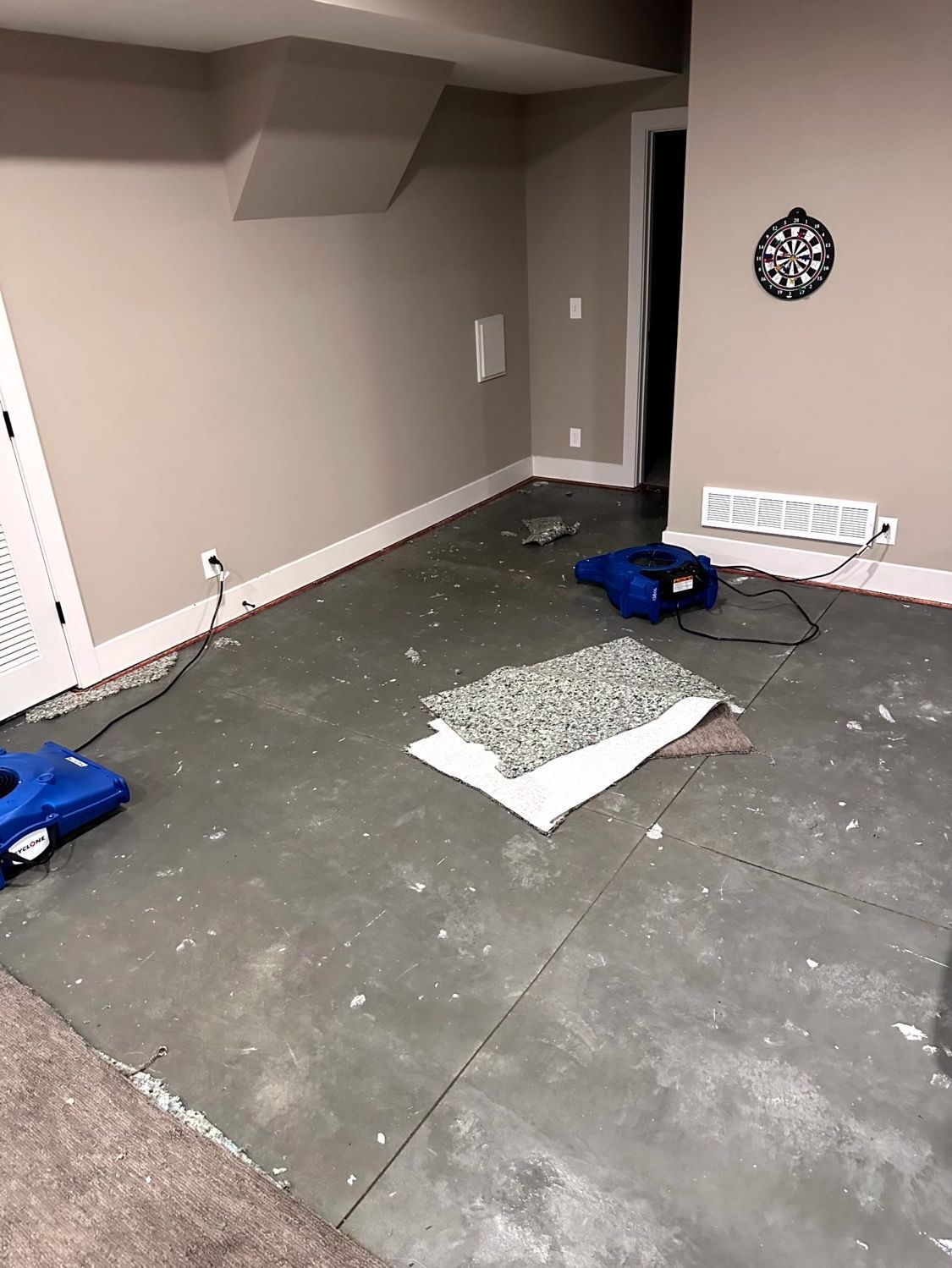 A basement undergoing water damage restoration, featuring bare concrete floors, a drying fan, and scattered debris.