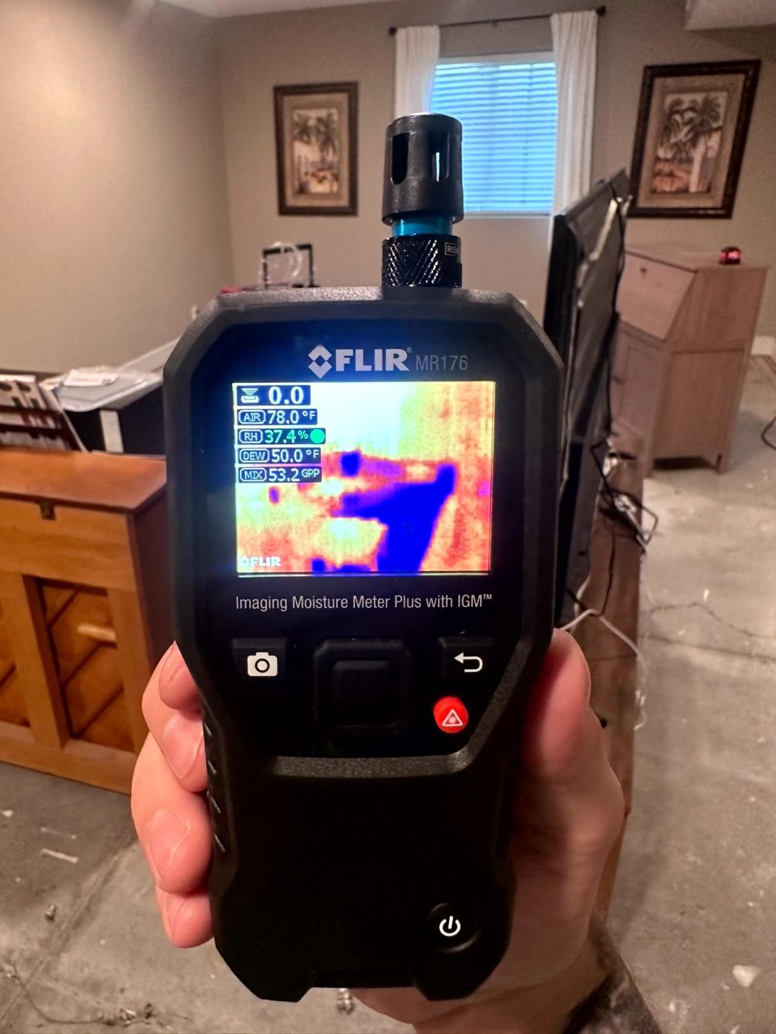 A hand holding a FLIR moisture meter displaying a thermal image of a wall, possibly detecting moisture or heat loss.