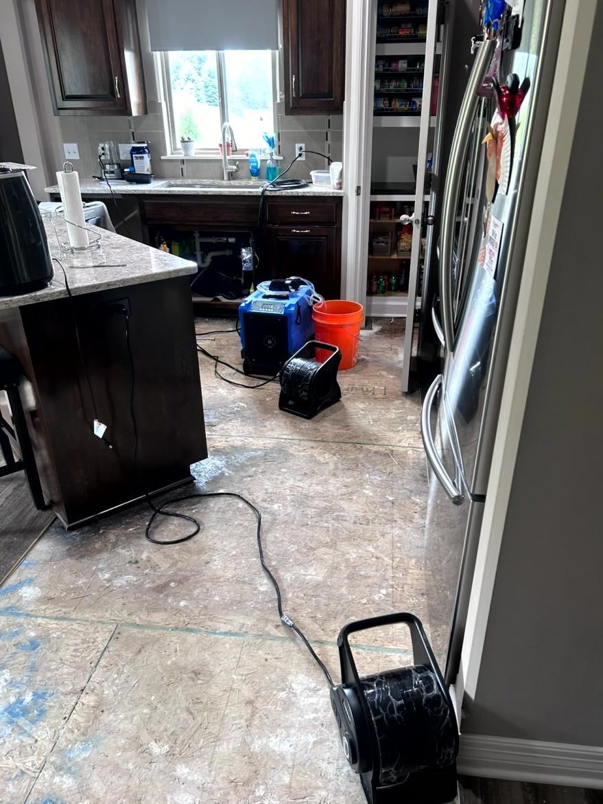 A kitchen floor with removed flooring, exposing the subfloor, with drying equipment and an orange bucket in the middle.