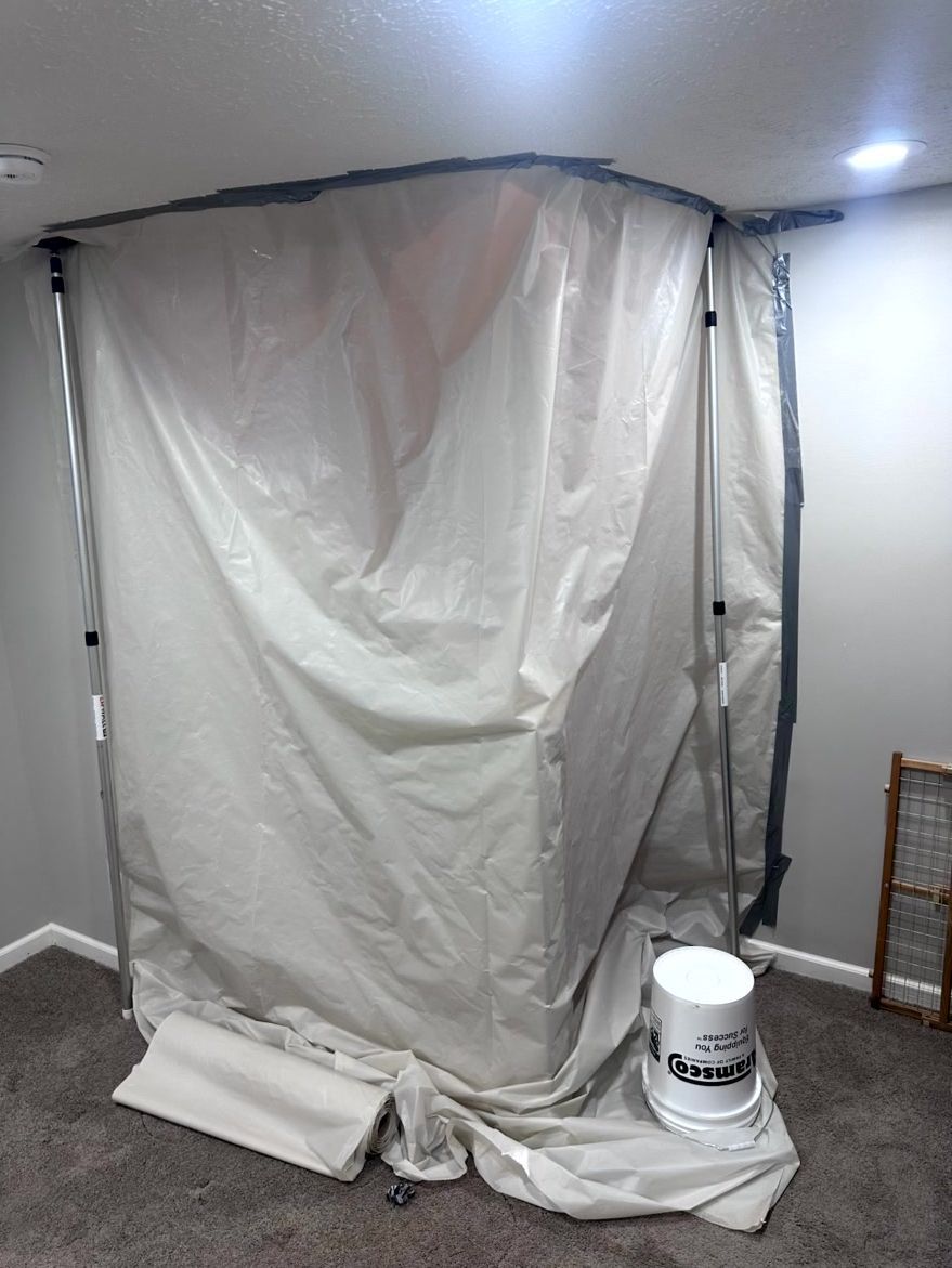 A temporary plastic dust barrier supported by poles stands in a carpeted room, with a bucket on the floor nearby.